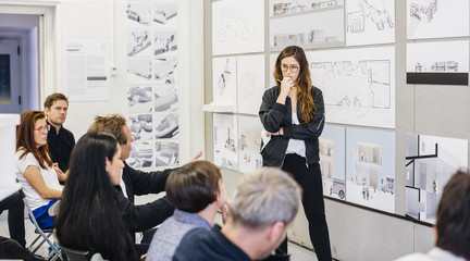A female student standing in front of a group of seating jurors evaluating the work mounted on the wall behind her