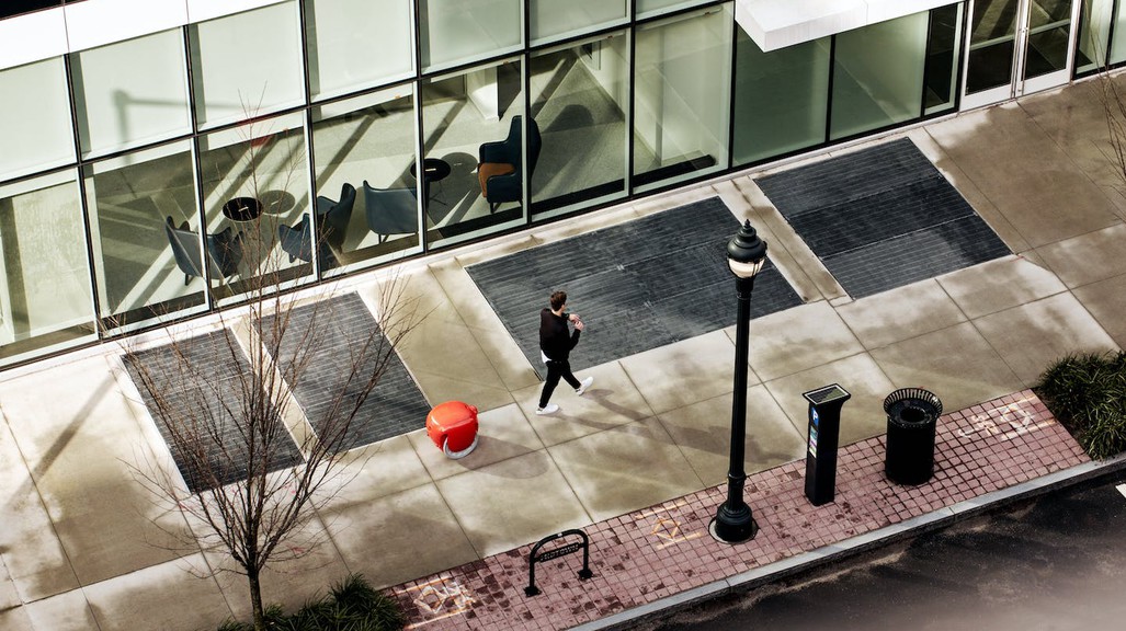 A photograph of a man walking down a city sidewalk with a small, orange robot following behind him