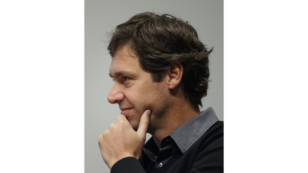 A photograph of a man with brown hair, with his hand on his chin, and smiling