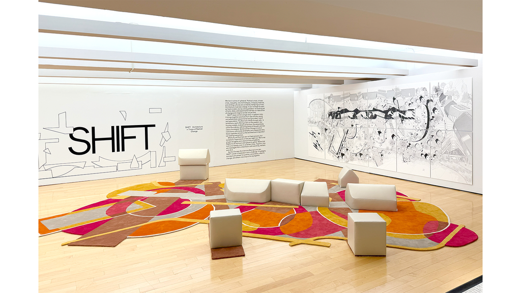 A bright photo of a large gallery space with while walls and illustrations on the walls, and with a colorful carpet in the center of the room