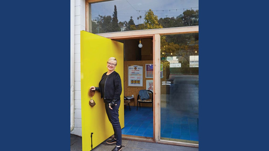 A woman standing in a doorway with a yellow door and a blue background, as she smiles toward the camera