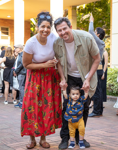A woman in a red dress and white shirt standing next to a man with a tan shirt, who is holding their baby