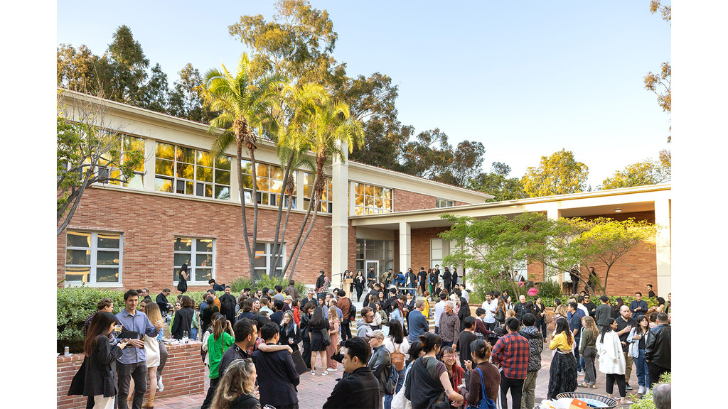 A crowd of people standing in an open courtyard, with a brick building in the background and a mix of blue sky and green trees