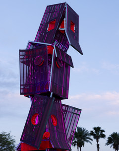 A photograph of a tall structure made of different, red-colored panels and screens, placed on a large open lawn area with a blue sky and people standing around the structure