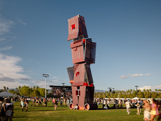 A photograph of a tall structure made of different, red-colored panels and screens, placed on a large open lawn area with a blue sky and people standing around the structure