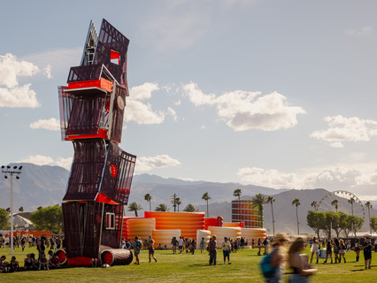 A photograph of a tall structure made of different, red-colored panels and screens, placed on a large open lawn area with a blue sky and people standing around the structure
