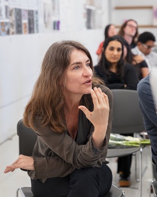 A woman with brown hair and wearing a tan shirt is sitting in a white classroom, gesturing with her right hand