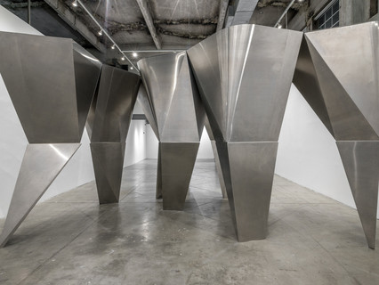 A photograph showing five silver, aluminum columns of different shapes and sizes, standing together in a loose row