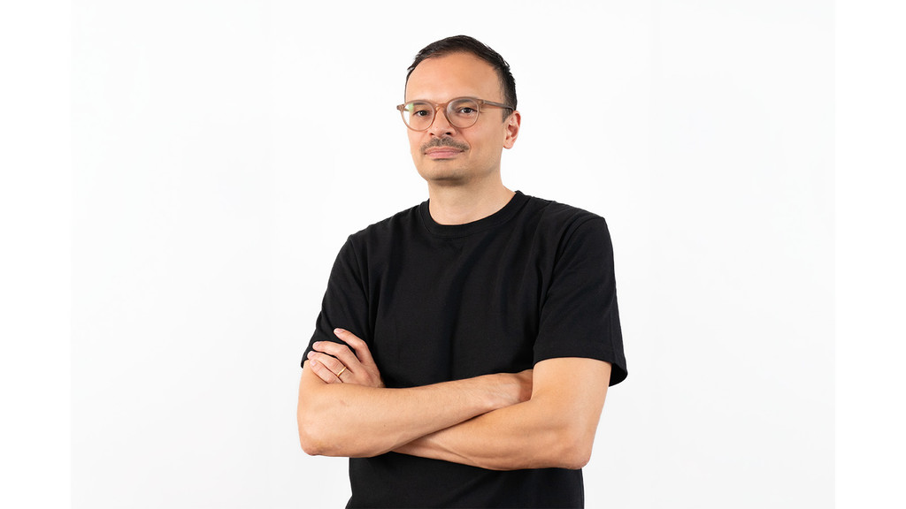 A man wearing a black t-shirt is standing with his arms crossed, wearing tan colored eyeglasses and smiling slightly toward the camera, with a plain white background