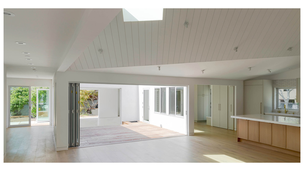 A photograph of the interior of a house, showing white walls and an angular white ceiling, with warm sunlight in some areas and some shadows in other areas, and a kitchen counter and sink in the background