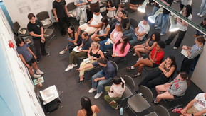 A photograph from above, showing students sitting in rows of chairs watching three other students present models of architectural work