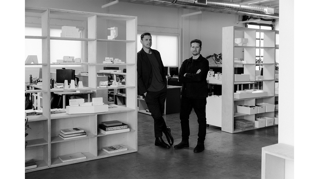 A black and white photo of two men standing between two bookcases, facing the camera, with various building models and tools on the shelves