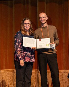 a photograph of a woman and a man standing on stage holding an award certificate